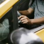 From above of crop unrecognizable male in polo shirt driving yellow cab on city street in daytime