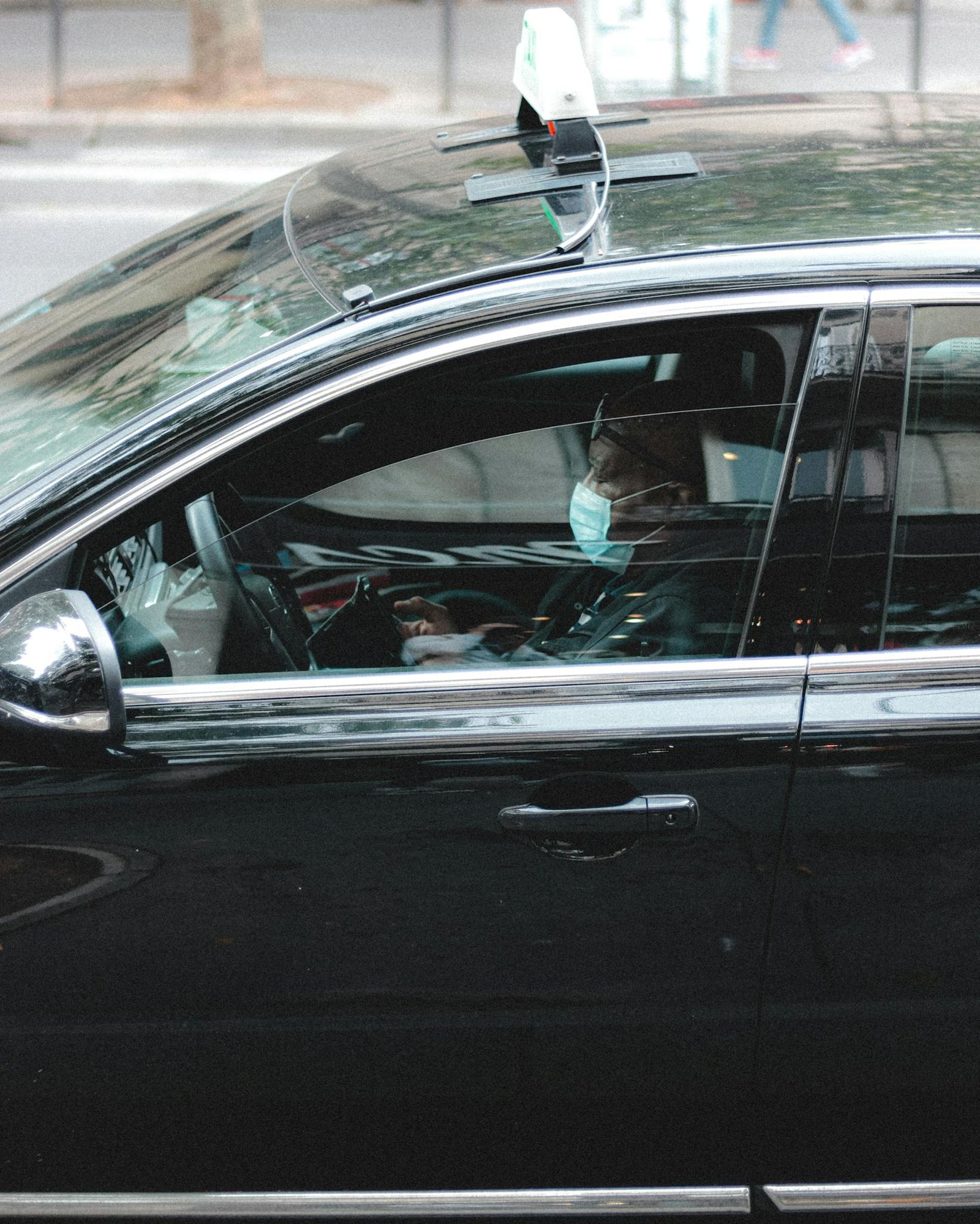 Side view of African American male driver in mask driving black taxi on road in downtown of big city at  daytime